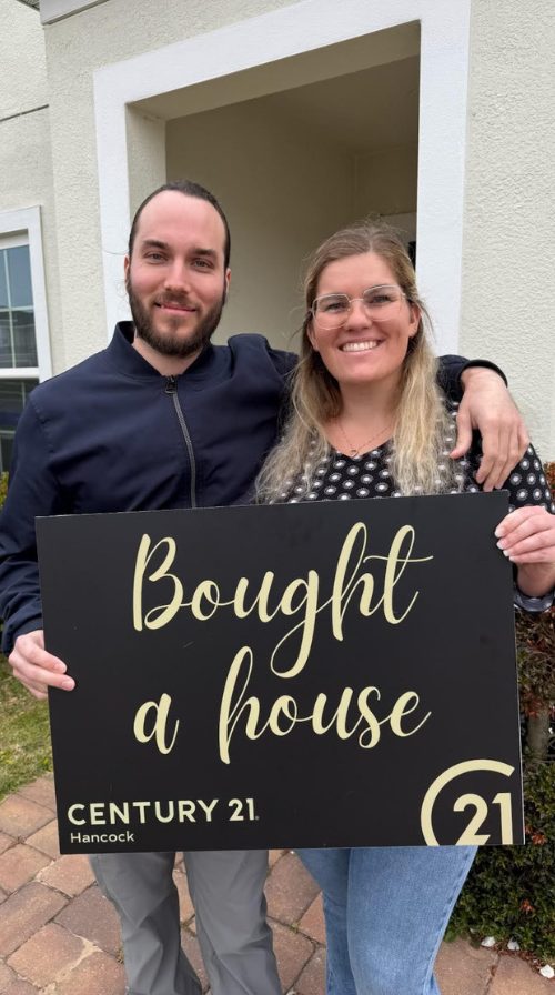 Couple holding a "Bought a house" sign outdoors next to a Florida home entry.